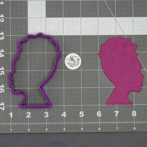 Puede incluir: Un cortador de galletas de plástico morado y una galleta morada con forma de perfil de mujer. El cortador de galletas está a la izquierda y la galleta a la derecha. Una moneda estadounidense está al lado del cortador de galletas para comparar el tamaño. El fondo es una esterilla de corte gris.