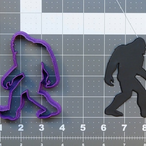Pode incluir: Cortador de biscoitos de plástico roxo em forma de Pé Grande caminhando. O cortador de biscoitos está em um fundo de grade cinza.