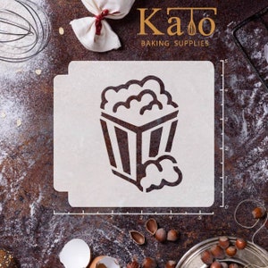 May include: A white plastic stencil with a black outline of a popcorn bucket with popcorn spilling out of the top. The stencil is 5 inches wide and 5 inches tall. The stencil is on a brown surface with flour and other baking supplies.