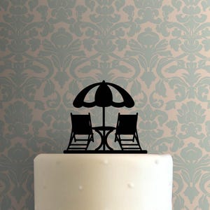 May include: Black silhouette cake topper featuring a beach scene with two lounge chairs, a small table, and a large umbrella. The topper is set against a patterned background, suggesting a vacation or summer theme.
