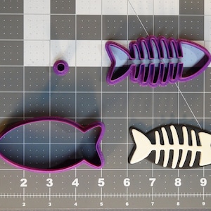 Könnte beinhalten: Drei violette Plastik-Ausstecher in Fischform mit Skelett-Design. Ein Ausstecher ist durchgehend, während die anderen beiden ein ausgeschnittenes Skelett-Design haben.
