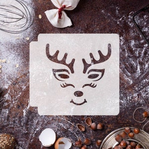 May include: A white plastic stencil with a reindeer face design. The stencil is on a brown surface with flour scattered around it.