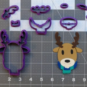 May include: A set of purple plastic cookie cutters in the shape of a reindeer, antlers, and other holiday-themed shapes. The cutters are on a gray grid background.