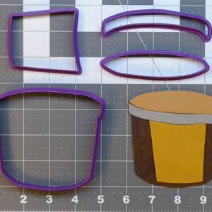 May include: A set of four purple plastic cookie cutters shaped like a drum, a cup, and two curved lines. The drum cookie cutter has a brown and yellow design.