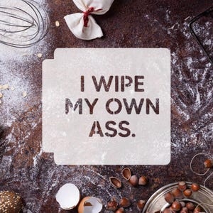 May include: A white stencil with the text "I WIPE MY OWN ASS" dusted with flour, alongside baking ingredients such as a whisk, hazelnuts, and an eggshell, on a brown surface.