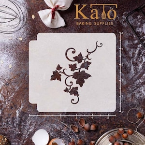 May include: A white plastic stencil featuring a brown ivy design. The stencil measures 12.7 cm wide and 12.7 cm tall. It is displayed on a brown surface with a ruler and other baking supplies.