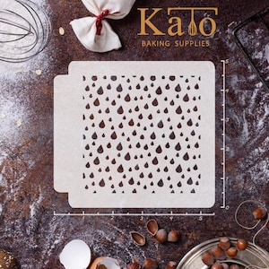 May include: A white plastic stencil with a repeating pattern of raindrops. The stencil is 5 inches wide and 5 inches tall. The stencil is on a brown surface with a ruler and other baking supplies.