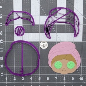 Pode incluir: Cortadores de biscoitos de plástico roxo em forma de rosto de mulher, cabelo e fatia de pepino. O rosto tem uma toalha rosa na cabeça e fatias de pepino verdes sobre os olhos.