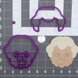 May include: Two purple plastic cookie cutters in the shape of a woman's face. One cutter has hair and the other has glasses.