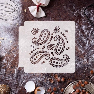 May include: White plastic stencil with a paisley design. The stencil is on a brown surface with flour scattered around it.