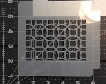 May include: A white plastic stencil featuring a geometric design of overlapping circles and squares. The stencil measures approximately 15 cm wide and 15 cm tall.