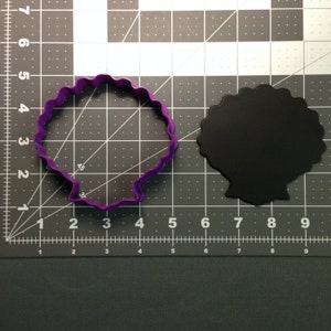 May include: Purple plastic cookie cutter shaped like a seashell on a gray grid surface.