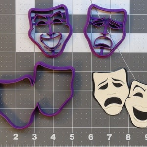 May include: Four purple plastic cookie cutters shaped like theater masks. Two cutters are of a smiling face and two cutters are of a frowning face.