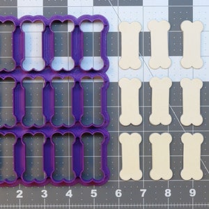 May include: A purple plastic cookie cutter with 12 bone-shaped cutouts. The cutter is on a grey grid background. There are 12 bone-shaped cookies next to the cutter.