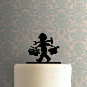 Op de afbeelding: Zwart silhouet taarttopper met een kind als bouwvakker met gereedschap. Het kind draagt een helm en draagt een moersleutel, hamer en gereedschapskisten. De topper staat op een witte taart met een patroon.