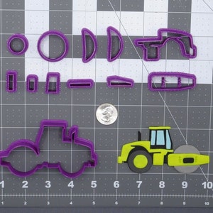 Op de afbeelding: Paarse plastic koekjesvormen in de vorm van een wegwals, een tractor en verschillende andere vormen. Een gele wegwals koekjesvorm is ook te zien.