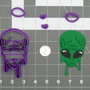 May include: A set of purple and green cookie cutters shaped like an alien head, dripping green goo, and a face with a striped pattern. The cutters are on a grey grid background.