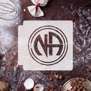 Puede incluir: Un esténcil blanco con las letras "NA" dentro de un círculo, probablemente para hornear o manualidades. El esténcil está sobre una superficie marrón espolvoreada con harina. Otros elementos de repostería son visibles.