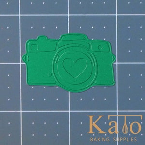 May include: Green metal cookie cutter shaped like a camera with a heart in the lens.
