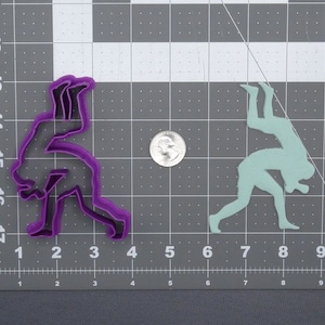May include: Purple and light blue plastic cookie cutters shaped like two people wrestling. The cutters are on a gray grid background.