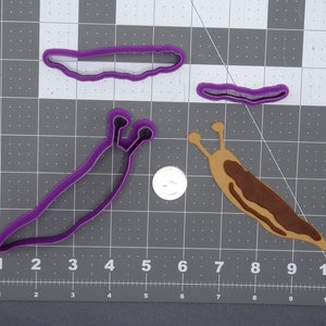May include: Three purple plastic cookie cutters shaped like slugs. One cutter is a simple slug shape, another is a slug with a curled tail, and the third is a slug with a curled tail and two antennae.