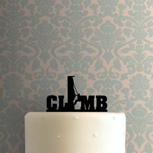 May include: A black cake topper with the word "CLIMB" and a silhouette of a person climbing a rock face. The topper is placed on a white cake, with a patterned background.