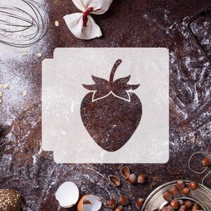 May include: A square stencil featuring a central cutout of a strawberry shape with a stem and leaves. The stencil is white and has a tab on the top edge. It is positioned against a dark, textured surface dusted with white powder and scattered nuts.