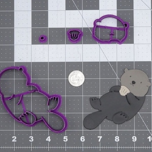 May include: Purple plastic cookie cutters in the shapes of a sea otter holding a fish, a sea otter, and a fish. The cutters are on a gray grid background.