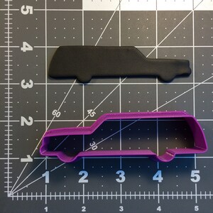 May include: A purple plastic cookie cutter in the shape of an estate car. The cookie cutter is on a grey gridded surface.
