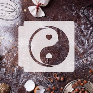 Puede incluir: Plantilla blanca de un símbolo yin yang con dos corazones en su interior. La plantilla está sobre una superficie marrón con harina y otros ingredientes para hornear.