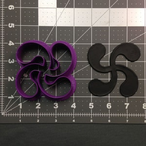 May include: A purple plastic cookie cutter shaped like a four-leaf clover with rounded edges. A black plastic cookie cutter of the same shape is shown to the right.