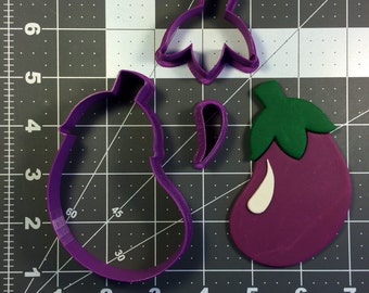 May include: Purple plastic cookie cutters in the shape of an eggplant, including the body, stem, and leaf. A finished eggplant cookie is also shown, with a purple body, green stem, and white accents.
