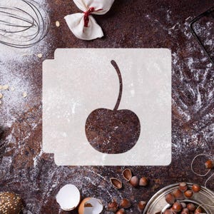 May include: A square white stencil featuring a centered cutout of a single cherry with a long stem. The stencil is positioned flat, displaying a clean, die-cut silhouette of the fruit shape, designed for creating decorative patterns on surfaces.