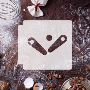 May include: A square white plastic stencil featuring a central cutout design of two angled bars and a small circle positioned between them. The stencil is placed on a dark, textured surface dusted with white flour, surrounded by baking tools.