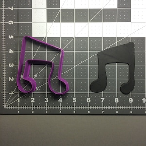 May include: A purple plastic music note cookie cutter on a gray grid surface. A black music note cookie cutter is to the right of the purple cutter.