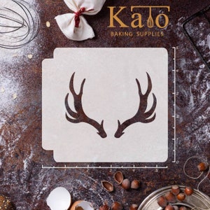 May include: A white plastic stencil with a silhouette of a set of antlers. The stencil is 5 inches wide and 4 inches tall. The stencil is on a brown surface with a ruler and other baking supplies.