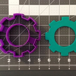 May include: Two plastic cookie cutters shaped like gears. The larger gear is green and the smaller gear is purple.