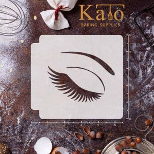 May include: A white stencil with a black outline of an eye with eyelashes. The stencil is 5 inches wide and 4 inches tall. The stencil is on a brown surface with a ruler.