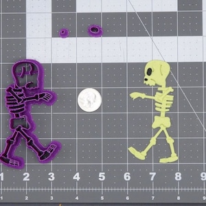 May include: Two plastic skeleton cookie cutters, one purple and one yellow, on a gray grid background. The purple skeleton is walking to the left and the yellow skeleton is walking to the right.