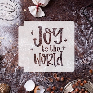 Puede incluir: Una plantilla blanca con el texto "Joy to the World" en una fuente decorativa. La plantilla está sobre una superficie marrón con harina esparcida alrededor.