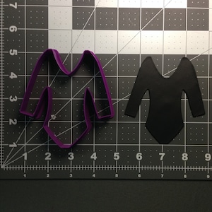 May include: Purple plastic cookie cutter shaped like a long-sleeved leotard. A black leotard cutout is next to the cookie cutter.