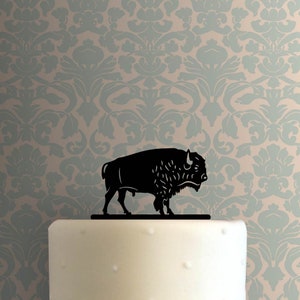 May include: A black bison cake topper. The silhouette of the bison is set against a white cake and a patterned wallpaper background. The bison is standing on a thin black base.
