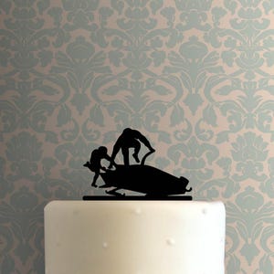 Puede incluir: Un adorno para pastel de silueta negra que representa a dos figuras empujando un bobsleigh. El adorno para pastel está colocado encima de un pastel blanco. El fondo es un patrón floral azul claro y beige.