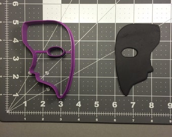 May include: Purple plastic cookie cutter shaped like a face mask with a single eye hole. A black plastic cookie cutter of the same design is shown to the right.
