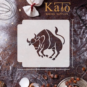 May include: A white plastic stencil of a bull, with a black silhouette, on a brown wooden surface. The stencil is 5 inches wide and 5 inches tall. The stencil is labeled "Kato Baking Supplies".