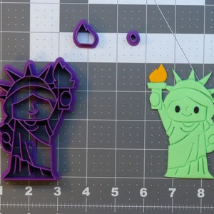 May include: Two cookie cutters, one purple and one green, shaped like the Statue of Liberty. The cutters are on a gray grid background.