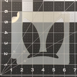 May include: A white plastic stencil with the outline of bunny ears. The stencil is approximately 6 inches wide and 4 inches tall.