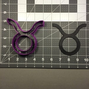 May include: Purple and black plastic cookie cutters shaped like the Taurus zodiac sign. The cutters are on a gray gridded surface.