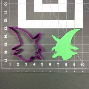 Puede incluir: Dos cortadores de galletas de plástico en forma de perfil de bruja. El cortador de la izquierda es morado y el de la derecha es verde. Ambos cortadores están sobre un fondo de rejilla gris.