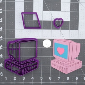 May include: Two purple plastic cookie cutters shaped like vintage computers. One computer has a heart on the screen and the other has a blank screen. There is also a small purple heart cookie cutter.